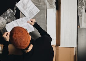 A person in a beanie reads an instruction manual while assembling furniture indoors.