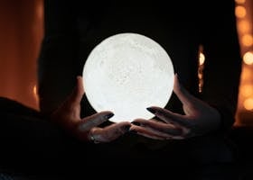 A person holding a glowing moon lamp, surrounded by warm bokeh lights, creating a mystical indoor atmosphere.