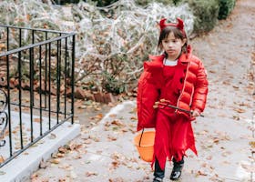 Little girl in red costume of devil for Halloween with horns and pitchfork walking on street in daytime in autumn