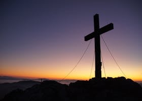 A dramatic cross stands silhouetted against a colorful sunset on a mountain peak, symbolizing faith and peace.